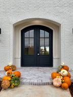 A front porch with a white brick wall and black double doors, decorated with orange, green, and white pumpkins on both sides of the entryway.