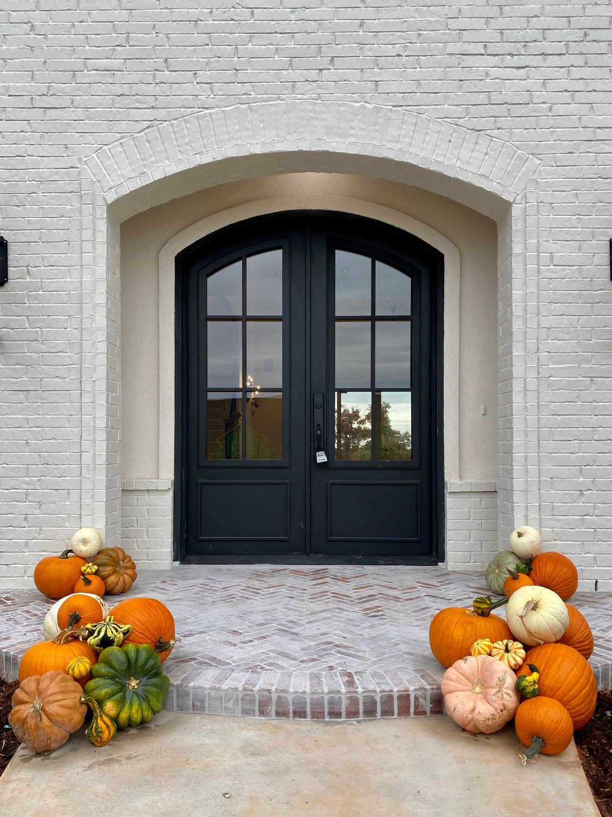 A front porch with a white brick wall and black double doors, decorated with orange, green, and white pumpkins on both sides of the entryway.